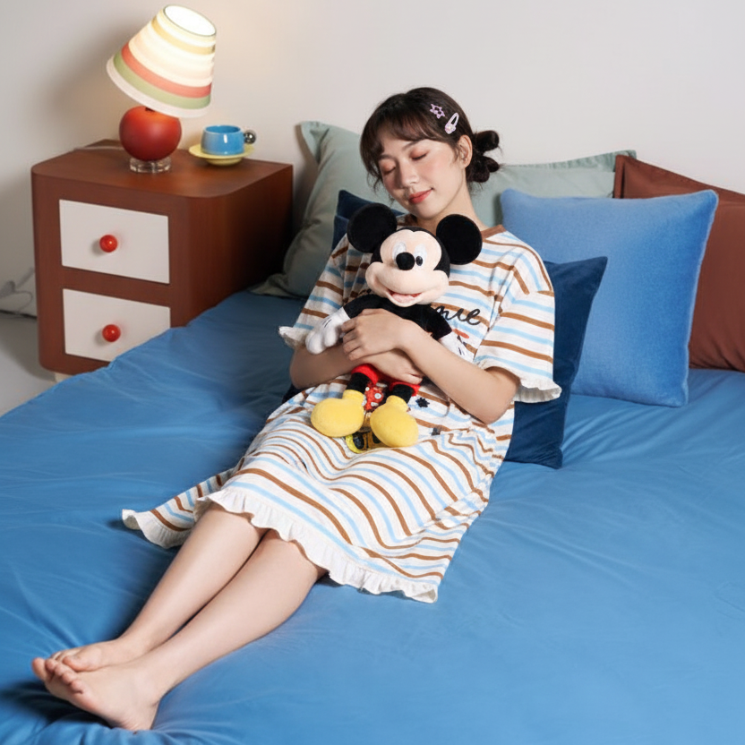 Woman sitting on a bed holding a plush toy, with a lamp and small table in the background.